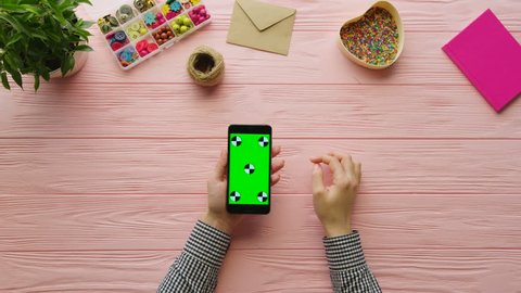 Woman using smart phone with green screen onpink wooden background. Female hands tapping on touch screen, scrolling. Chroma key, Tracking motion. Pink wooden desk with craft equipment . Top view Video de stock