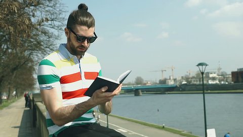 Handsome man in colorful shirt sitting on the wall next to the river and reading book
