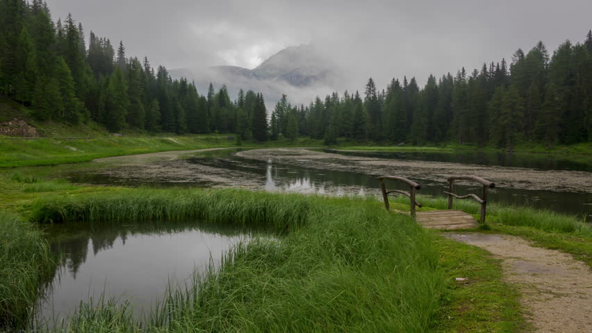 Summer evening. Forest pond and wooden bridge. Clouds and fog almost hiding the mountains in the background. Time lapse