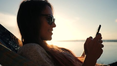 Young Smiling Mixed Race Tourist Girl in Sunglasses Using Mobile Phone in Hammock at the Beach near the Sea with Beautiful Sunset on Background. Koh Phangan, Thailand. HD Slowmotion.