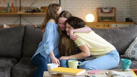 Two young woman bear up and calm down their best unhappy friend who is upset and depressed because of trouble and is needed a moral support, sitting on sofa at home during sunny day