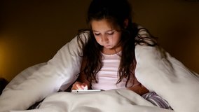 Cute little girl using tablet pc under blanket in a dark room. Winning gesture.