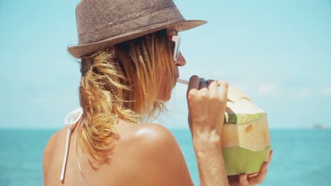 Woman drinking fresh coconut water with straw on beach fun vacation. Closeup of woman holding young green tropical fruit sipping for healthy snack during summer holidays in slow motion