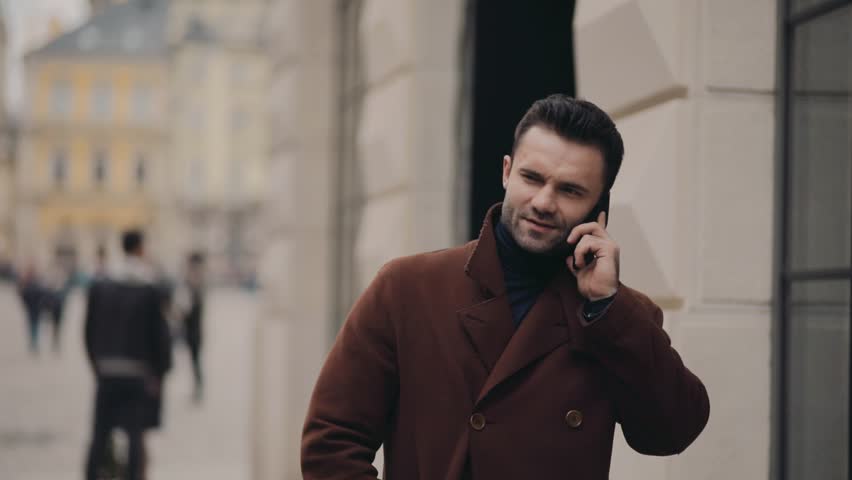 Attractive young man in a stylish coat walking down the crowded city street, the phone rings, he picks up the phone, happily talks, nodes, puts down the phone and continues his walk. | Shutterstock HD Video #26157119