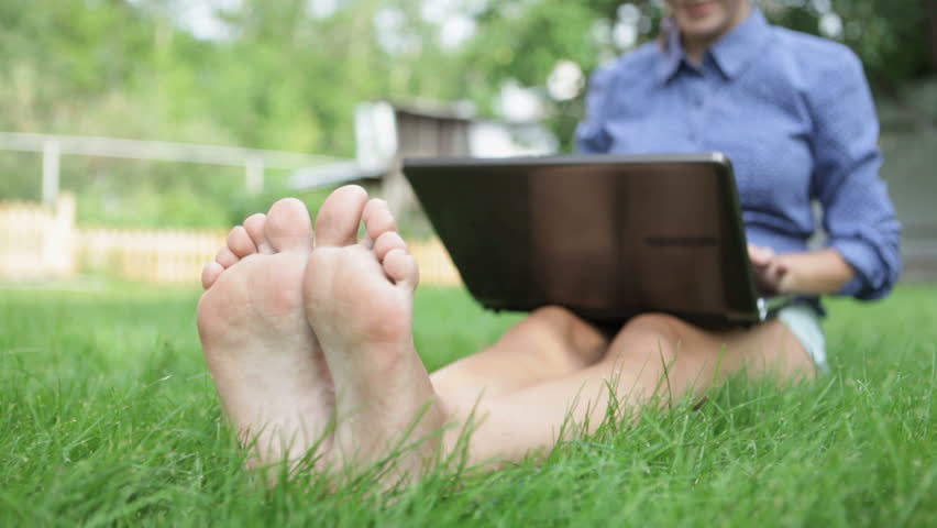 Close-up of barefoot woman sitting on the grass and typing letter on the laptop