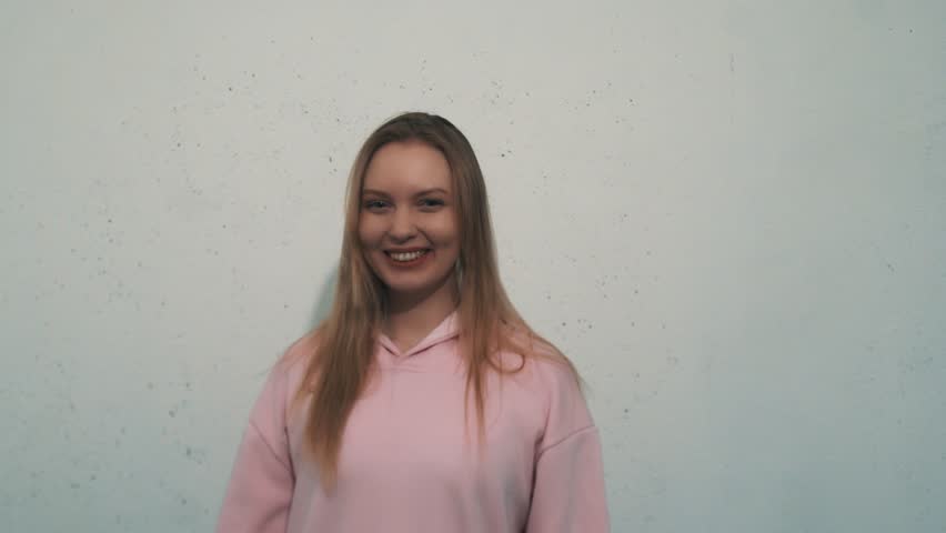 Vigorous young blonde caucasian girl with ear flesh tunnels in pink sweatshirt jumping around in excitement in front of white wall