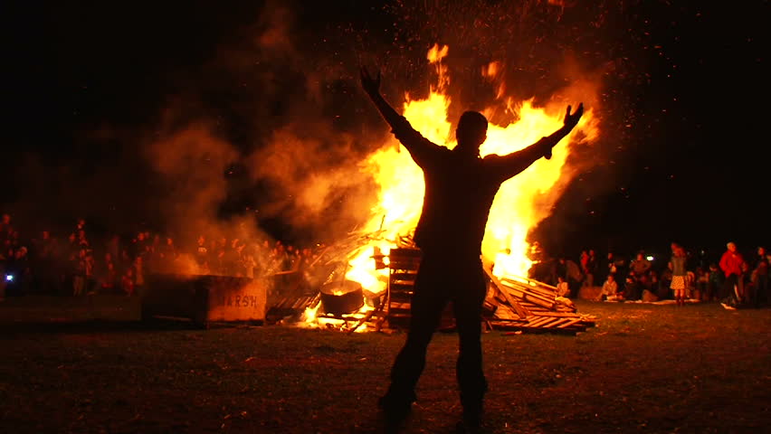 Man in front of large bonfire has both arms up high taking in the heat, filmed