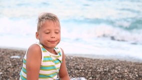 Child sits on stony beach and looks at camera at beginning of video then turns away to sea. Closeup portrait of cute blond boy isolated at beautiful blue sea background. Real time video footage.