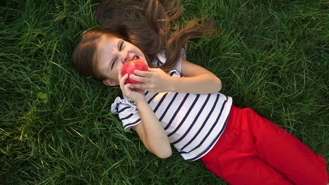 Little girl is eating an apple lying on the green grass in the park. Have a bite in the open air.
