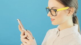 Brunette with Mobile Device Uses Touchscreen on Blue Background in Studio. Attractive Hipster Girl Wearing Fashionable Glasses and Using her Smartphone to Find Information on the Internet.