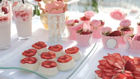 Sugary appetizers and strawberries on white table outdoor. Light jelly decorated with fresh cut berries, cookies on sticks in carved vases, creamy cheesecakes and strawberries spread out over paper