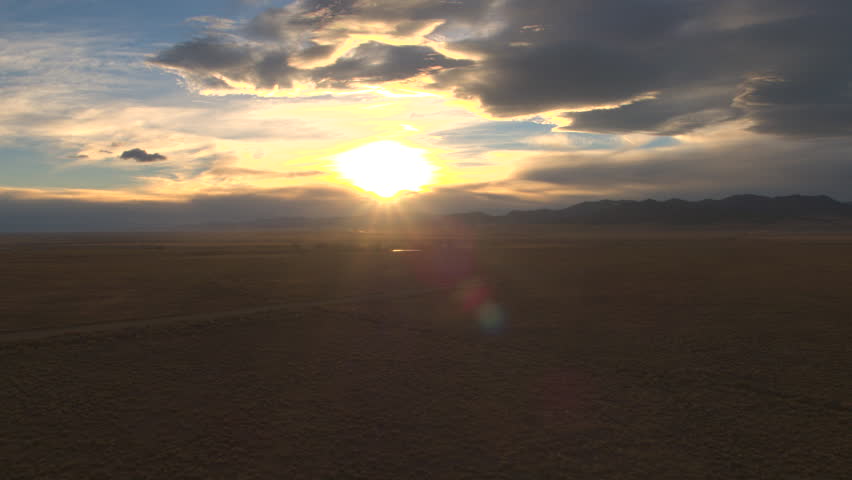 AERIAL: Flying above black SUV car driving through the desert valley at dramatic sunset. Wind blowing raised dust particles on the dirt track. People on road trip traveling to the Rocky Mountains
