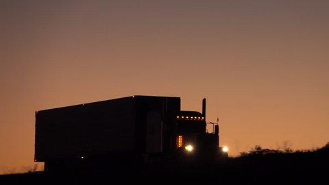 SLOW MOTION, CLOSE UP: Silhouetted freight container semi trailer trick traveling along the empty highway shipping cargo at peach-pink light of dawn. Transporter driving on the road hauling heavy load