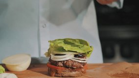 Male cooking chief in white robe finishes making hamburger and takes it in hands, sauce dripping on wooden cutting board
