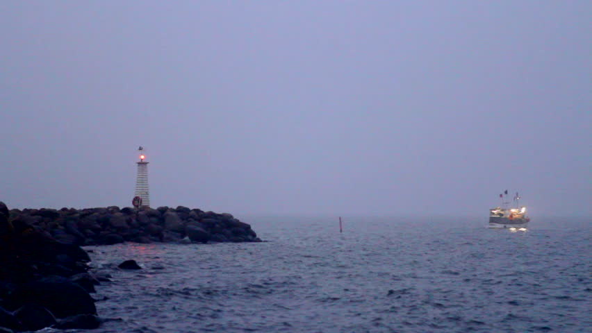 Wide shot of a lighthouse on a jetty with a boat sailing next to it at dusk