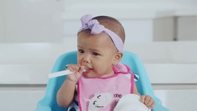 Video footage of a cute baby girl eating food in the kitchen while holding a spoon and bowl