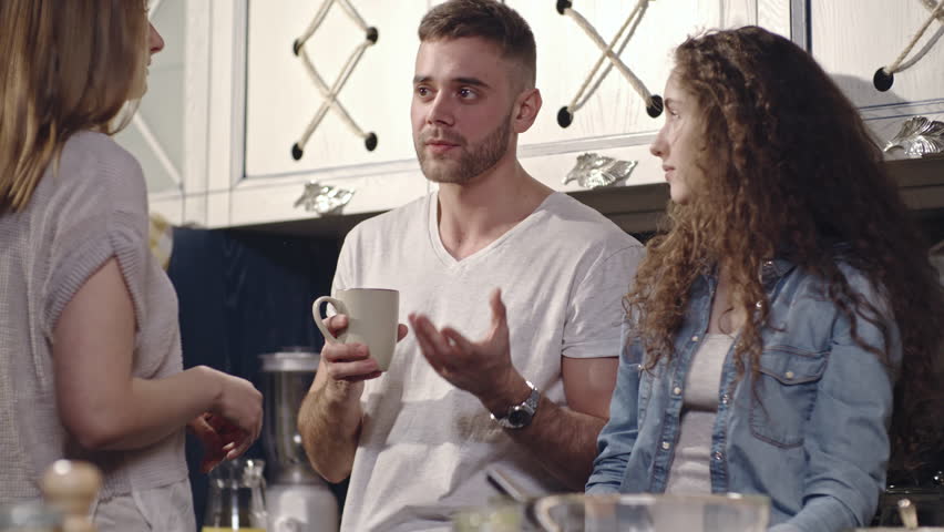 Tracking shot of two young women and man discussing something in kitchen and drinking tea