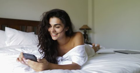 Young Woman Using Cell Smart Phone Happy Smiling Beautiful Girl Lying On Bed In Bedroom Morning Slow Motion 60