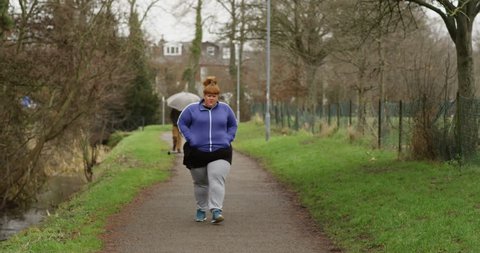 4K Unhappy overweight woman in leisure wear walking alone outdoors in the park. Social issues & unhealthy lifestyle concept. Slow motion.