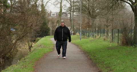 4K Unhappy overweight man walking towards camera, alone outdoors in the park. Social issues & unhealthy lifestyle concept. Slow motion.