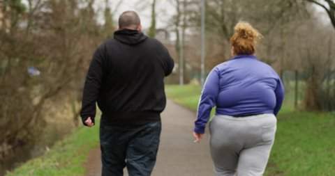 4K Obese couple walking away from camera, smoking cigarettes outdoors in the park. Social issues, unemployment & unhealthy lifestyle concept. Slow motion.