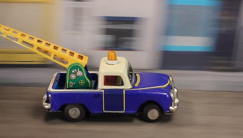 Tin toy breakdown truck towing a car along a urban road with emergency light flashing.