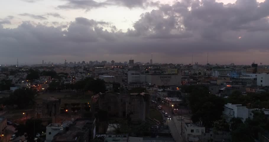 Descending aerial view toward a Santo Domingo street beneath dark clouds with the city skyline behind, 2017.