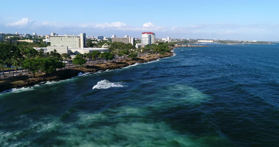 Aerial zoom toward a rocky Dominican Republic shoreline with clear Caribbean water, beachfront buildings, and distant city skyline, 2017.