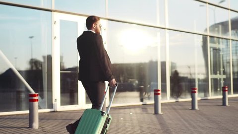 Young stylish bearded man in a suit walking to the airport, pulling suitcase and answering his phone call. Business style, active lifestyle. Traveling time.