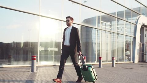 Stylish young bearded man in sunglasses exiting the airport terminal with luggage, answers the phone. Business style, traveler, modern lifestyle. Active lifestyle.