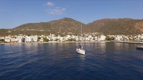 Sunny Morning in an empty Greek marina, Methana, Mediterranean Sea. Aerial video shooting, the height of a bird's flight. A lone sailing yacht leaves the marina. In the background is a landscape of