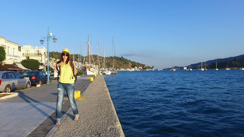 Tourist beautiful, young girl brunette in sunglasses in a yellow T-shirt, blue jeans, a yellow hat and with a colored backpack walks along the waterfront, at sunset, in sunlight, radiance, in the