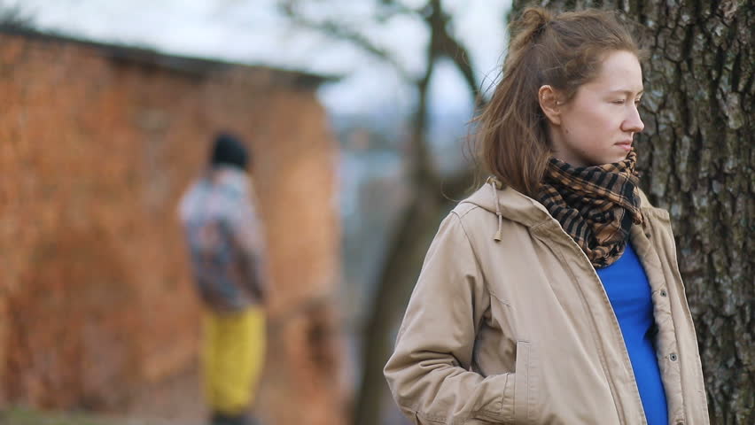Pretty youthful brown-haired pregnant girl in beige coat meeting sights with tall stranger in hood and yellow trousers on countryside landscape, focusing above them