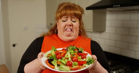 4K Portrait of fun overweight woman feeling sick looking at plate of salad, humorous dieting concept. Slow motion.