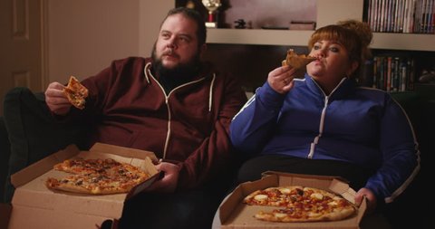 4K Overweight couch potato couple eating takeaway pizza in front of the TV