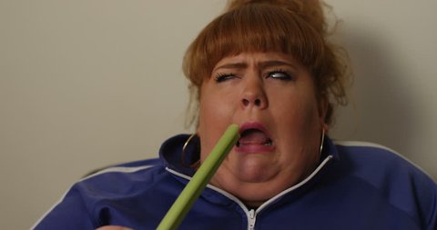 4K Unhappy overweight woman on a diet trying to force herself to eat a stick of celery. Funny dieting concept.