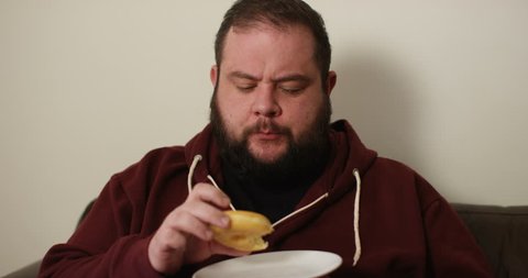 4K Happy obese man being a couch potato, eating a donut & really enjoying it