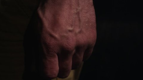 Hand of angry man in yellow pants with swollen veins clenches fist in black semi dark room, close up shot