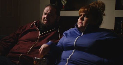 4K Funny couch potato couple eating junk food in front of the TV & reacting with horror