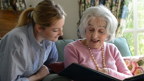 Senior woman sitting with adult granddaughter at home looking through photo album together