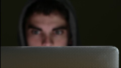 Focus is pulled from laptop to face of suspicious looking young man wearing hooded top working on laptop at night