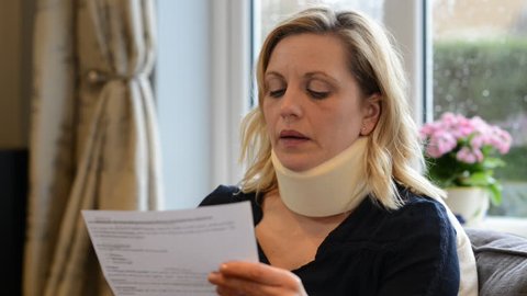 Mature woman at home wearing neck brace reads letter in respect of compensation for her injury - she winces in pain