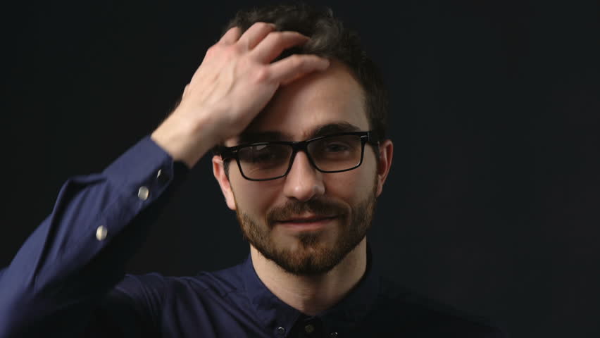 Closeup portrait of smiling young business wearing eyeglasses running hand through hair and smiling at camera, locked down video over dark background
