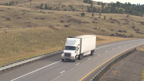 AERIAL CLOSE UP: White semi tuck transporting goods across the country in Great Plains, USA. Container freighter driving on empty interstate highway along the fields transporting cargo to the Rockies