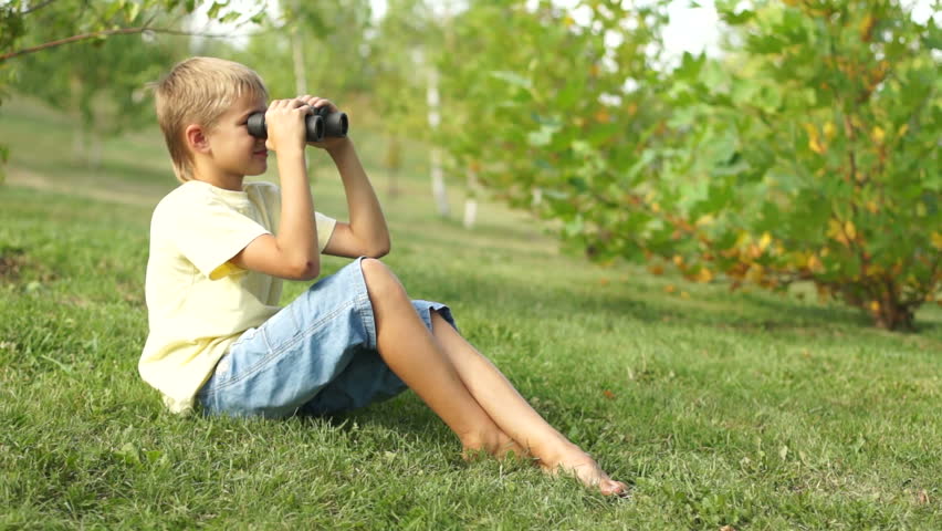 Boy watching with binoculars sitting on the grass. Thumbs up. Ok.
