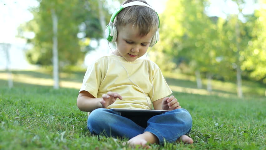 Cute little boy with headphones and a Tablet PC sitting on the grass. Online
