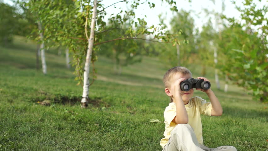 Little boy with binoculars sitting on the grass outdoors. Thumbs up. Ok.
