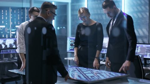 Team of Government Agents Tracking Fugitive with Help of Touchscreen Interactive Table in Big Monitoring Room Full of Computers with Animated Screens. Shot on RED EPIC-W 8K Helium Cinema Camera.