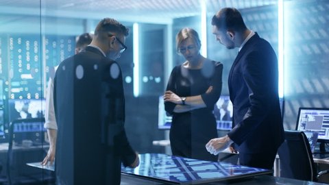 Team of Government Investigators Tracing Criminal with Help of Touchscreen Interactive Table in Big Monitoring Room Full of Computers with Animated Screens.