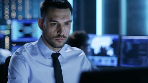 Close-up of a Professional Technical Controller Sitting at His Desk with Multiple Displays Before Him. In the Background His Colleagues Working in System Control Center.  Shot on RED EPIC-W 8K Helium 
